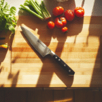 Top-down view of a kitchen countertop with a chef’s knife laid flat on a whetstone, surrounded by sharpening tools and fresh vegetables, brightly lit by natural window light in a modern rustic kitchen.