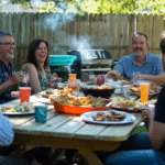 Realistic backyard BBQ with people eating at a picnic table, grillmaster cooking in the background