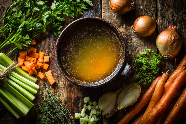 A steaming bowl of homemade golden vegetable broth recipe on a rustic wooden table, surrounded by fresh, colorful chopped vegetables like carrots, celery, and onions.