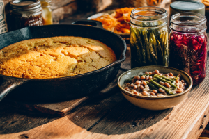 A rustic table showing Appalachian foods: a cast-iron skillet of cornbread, a bowl of soup beans, and pickled vegetables.