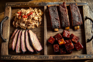 A wooden board showing four major BBQ styles: Carolina pulled pork, Memphis ribs, Texas brisket, and KC burnt ends.