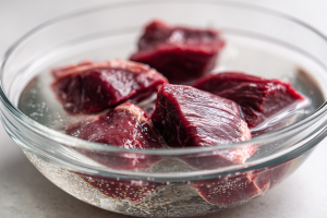 Pieces of raw, trimmed possum meat soaking in a large glass bowl of clear, salted water, a key step for removing gamey flavor.